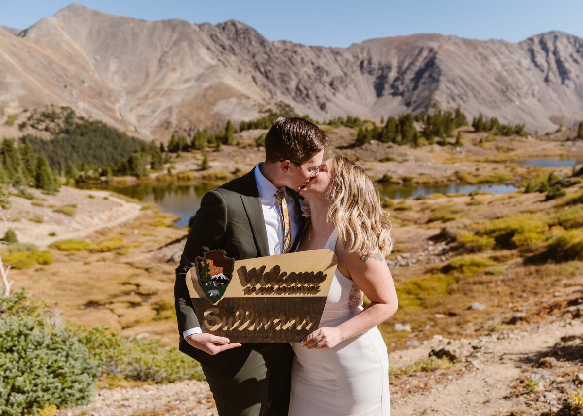 Two brides share a kiss while holding a custom wooden sign, with golden alpine scenery and mountain peaks in the background during their fall elopement in Keystone, Colorado.