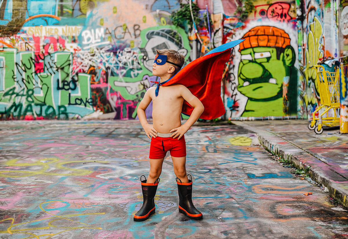 Little boy in red underwear and black rainboots wearing a superhero cape and mask in graffiti alley near MICA in Baltimore Maryland