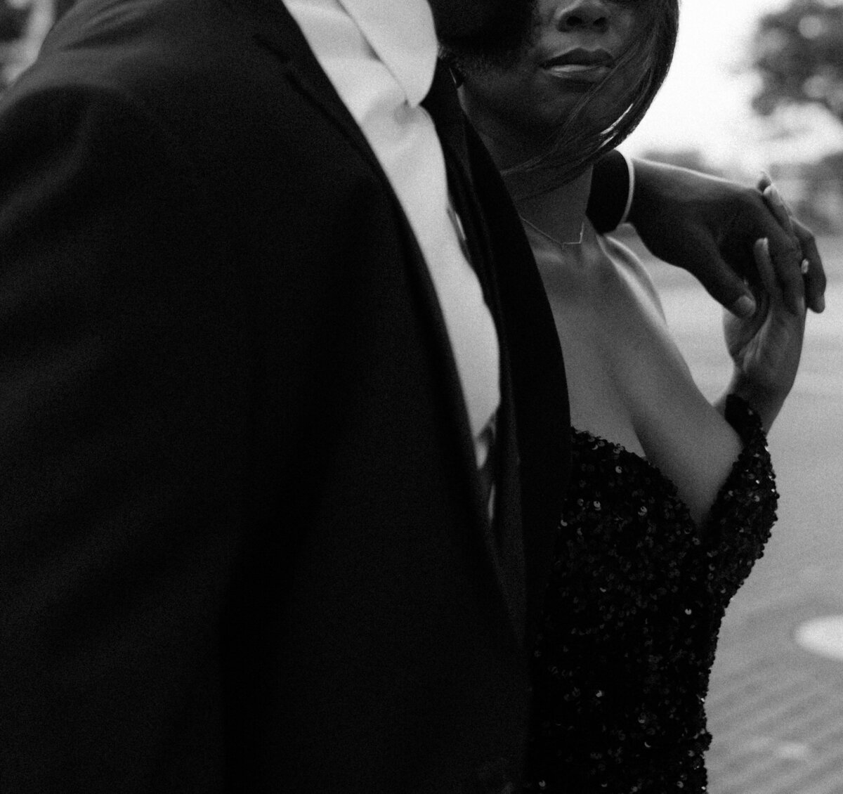 A black couple, photographed in black & white, in formal attire. The man has his arm around the woman, and they're holding hands