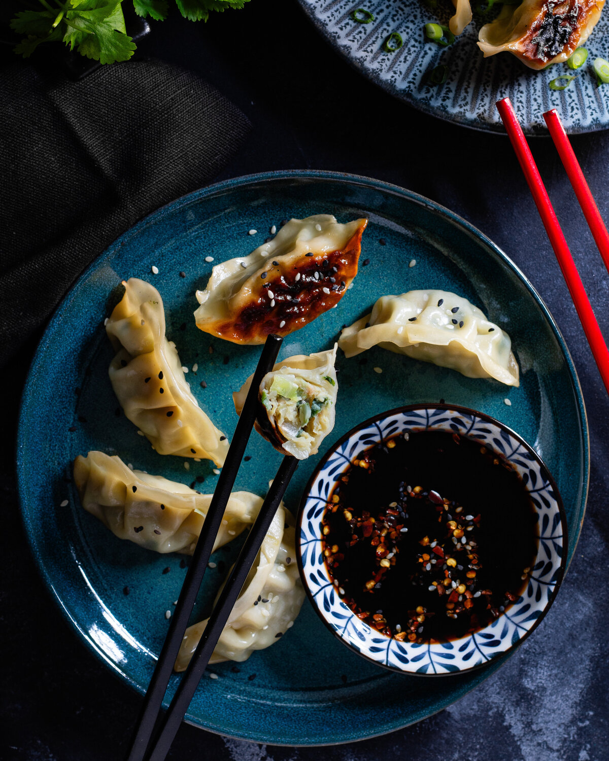 Chicken and vegetable gyoza  held by chopsticks with a plate of gyoza and dipping sauce