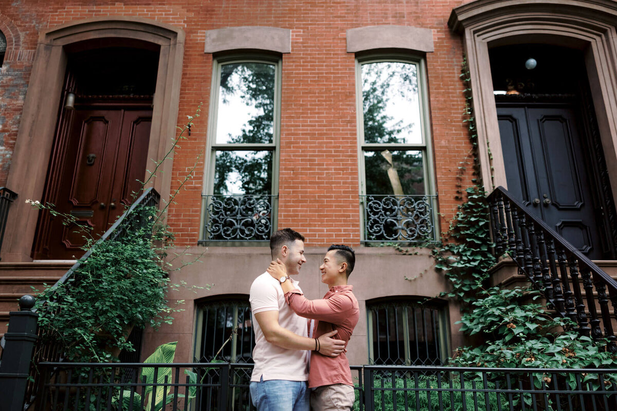 The engaged couple is happily staring at each other, standing close, in front of an ivy-covered building at West Village, NYC.