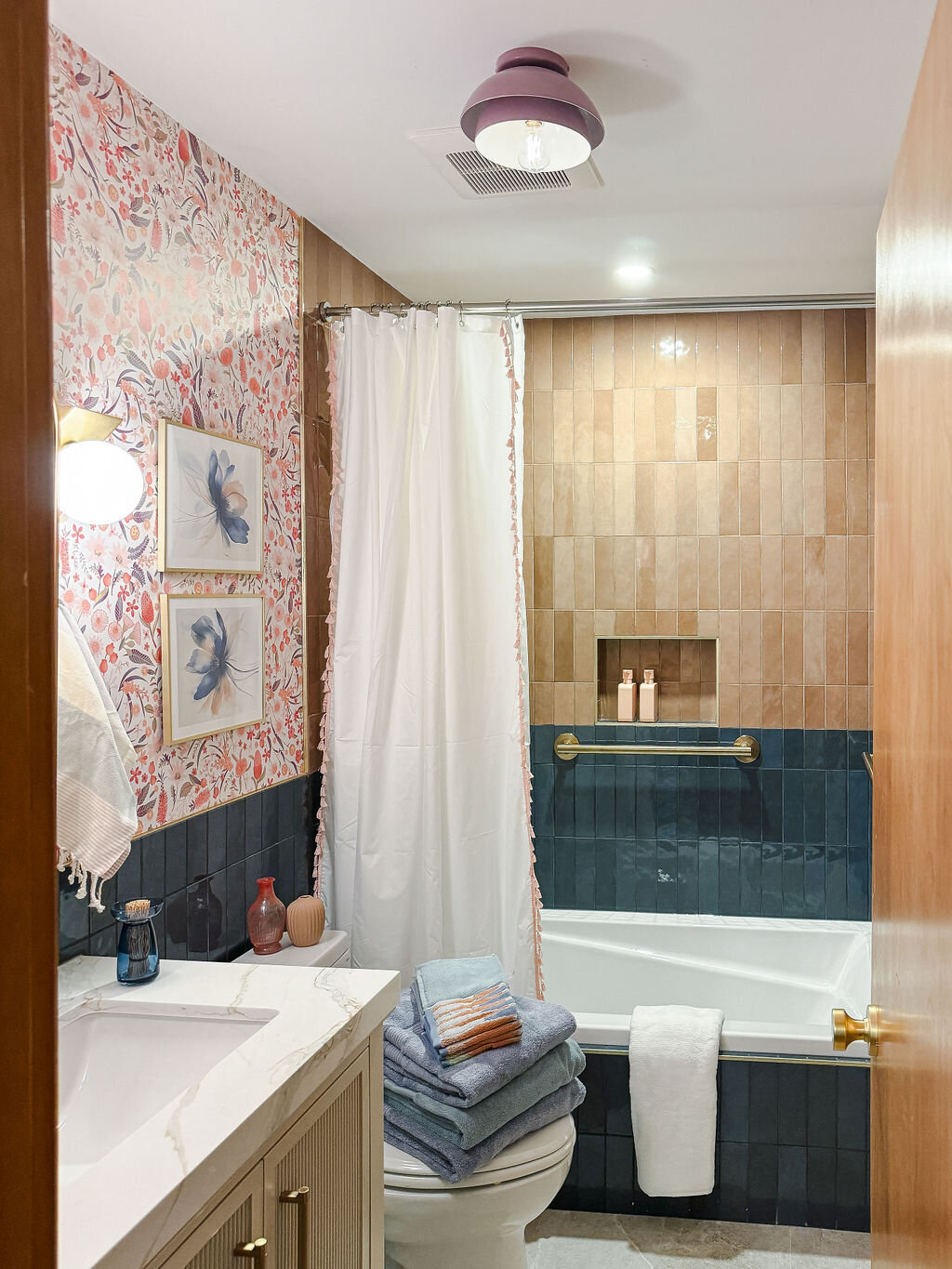 A brightly lit bathroom featuring a combination of floral wallpaper, warm-toned vertical tiles, and dark blue vertical tiles. A white bathtub with a brushed gold grab bar is partially visible, with a stack of folded blue towels on the toilet seat beside it and a white towel hanging over the edge. A white shower curtain with a delicate pink trim is drawn along a curved rod. A vanity with a white countertop and a blue glass bottle on it is visible on the left. Two framed floral watercolor prints hang on the wallpapered wall above the vanity. A recessed niche in the warm-toned tiled wall holds small toiletries. A modern ceiling light with a pink trim illuminates the room. A partially open wooden door is on the right.