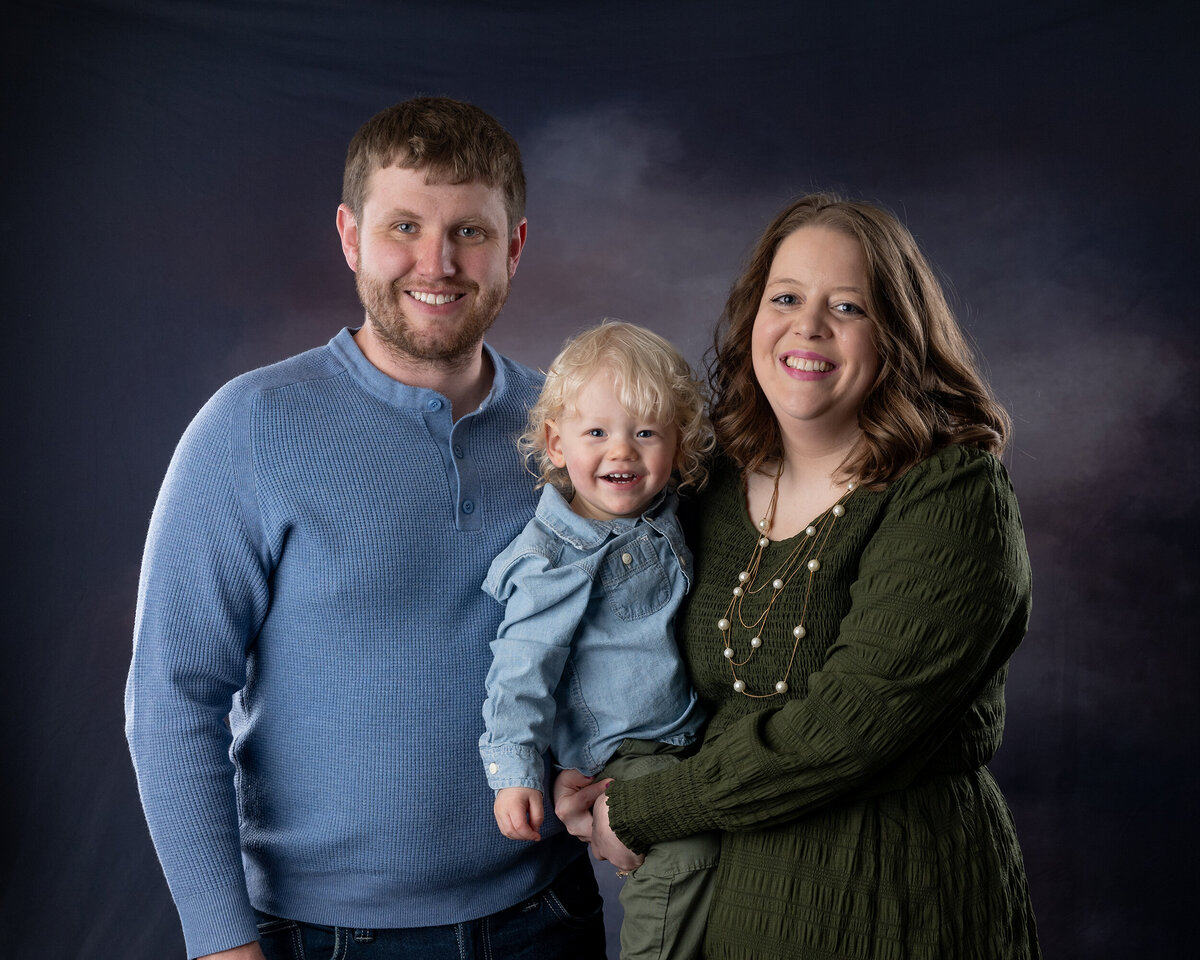 A smiling family, consisting of a man, a woman, and a young child with blonde curly hair, poses against a dark backdrop.