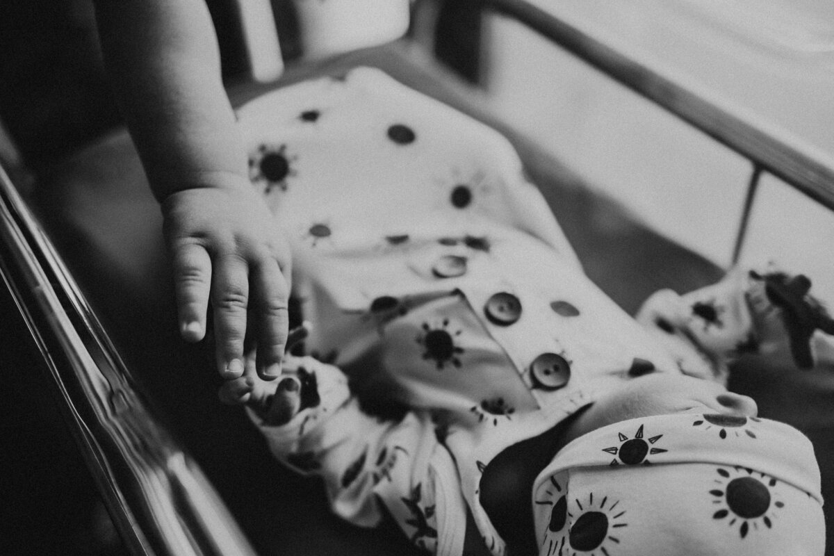 Albuquerque maternity photography studio captures a heartwarming scene with a newborn peacefully sleeping in his crib. His older sibling gently touches his hand, creating a touching moment of sibling connection and love.