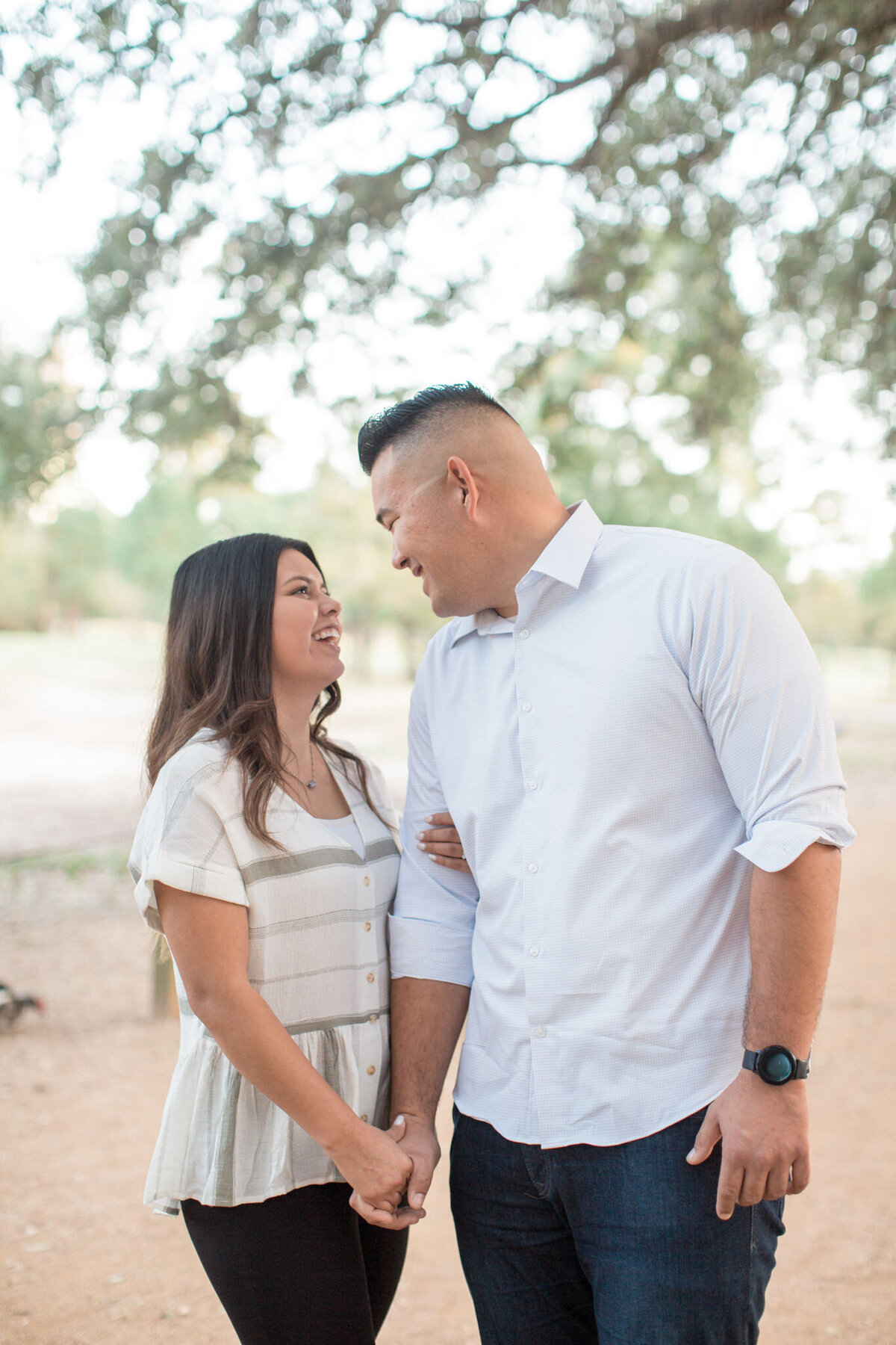 A couple stand on a path, her hand around his arm, smiling at each other and framed by oak limbs