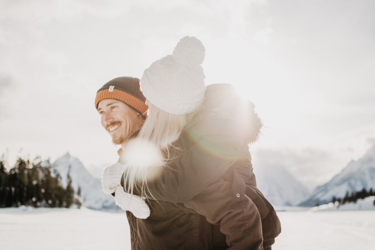 jackson hole photographers captures winter engagement with man smiling while holding his fiance on his back as the sun beaming through the woman's hair