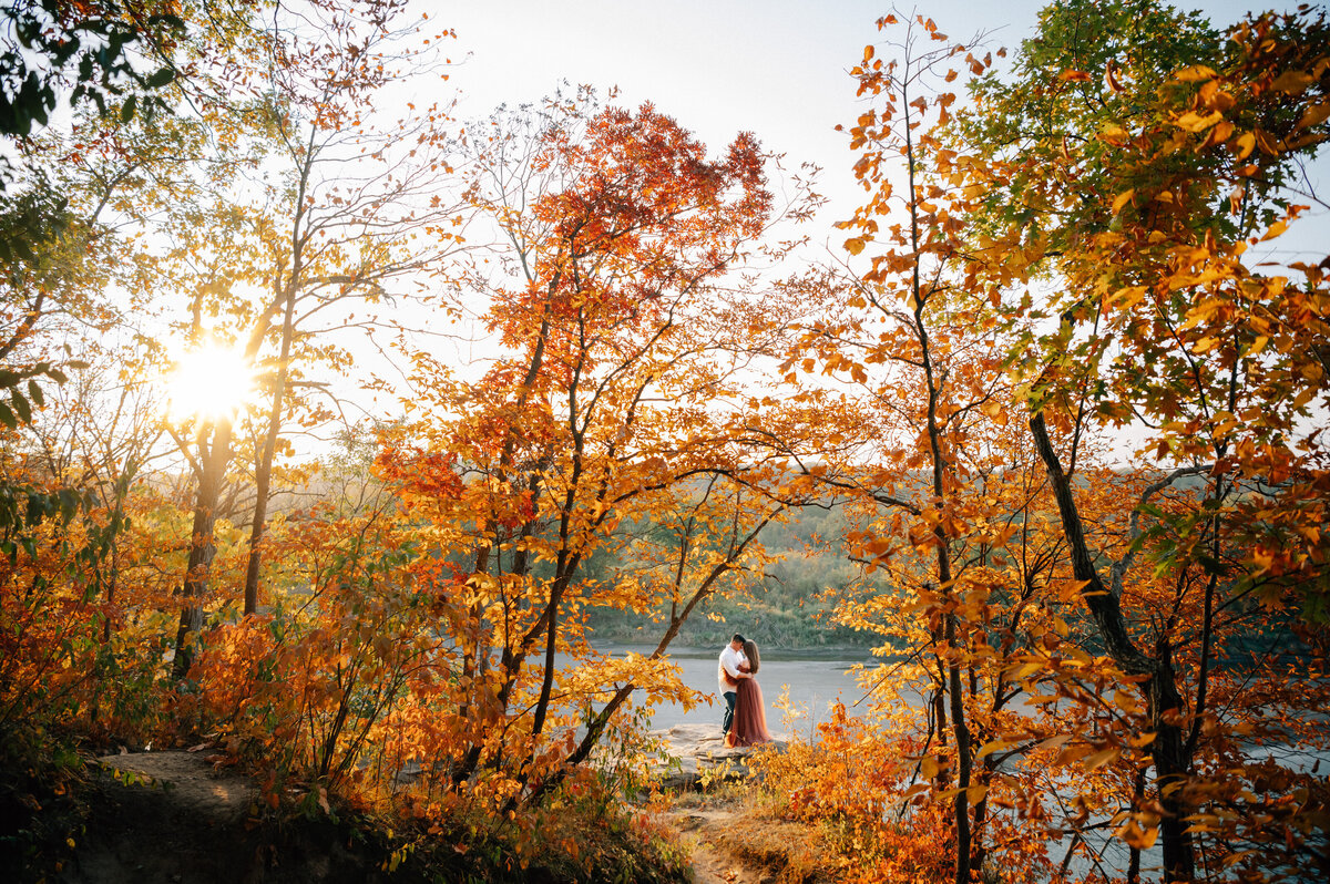 Engaged couple stands on a rock at Ledges State park surounded by fall trees while the sun peeks out through the trees. Photo by Anna Brace, a wedding photographer in Omaha.