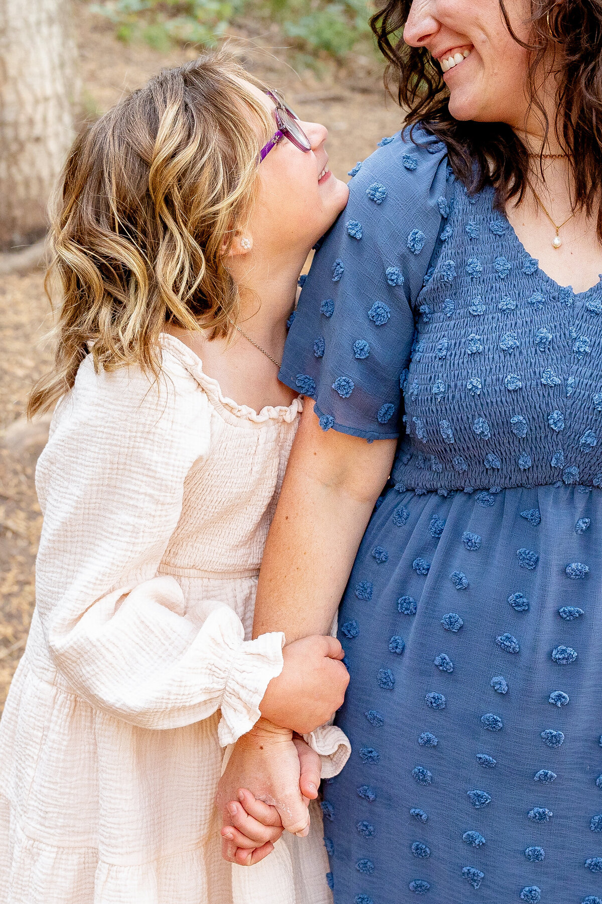 A young girl with blonde curls and purple glasses leans on her mother's shoulder, gazing up at her lovingly while holding hands in a natural outdoor setting.