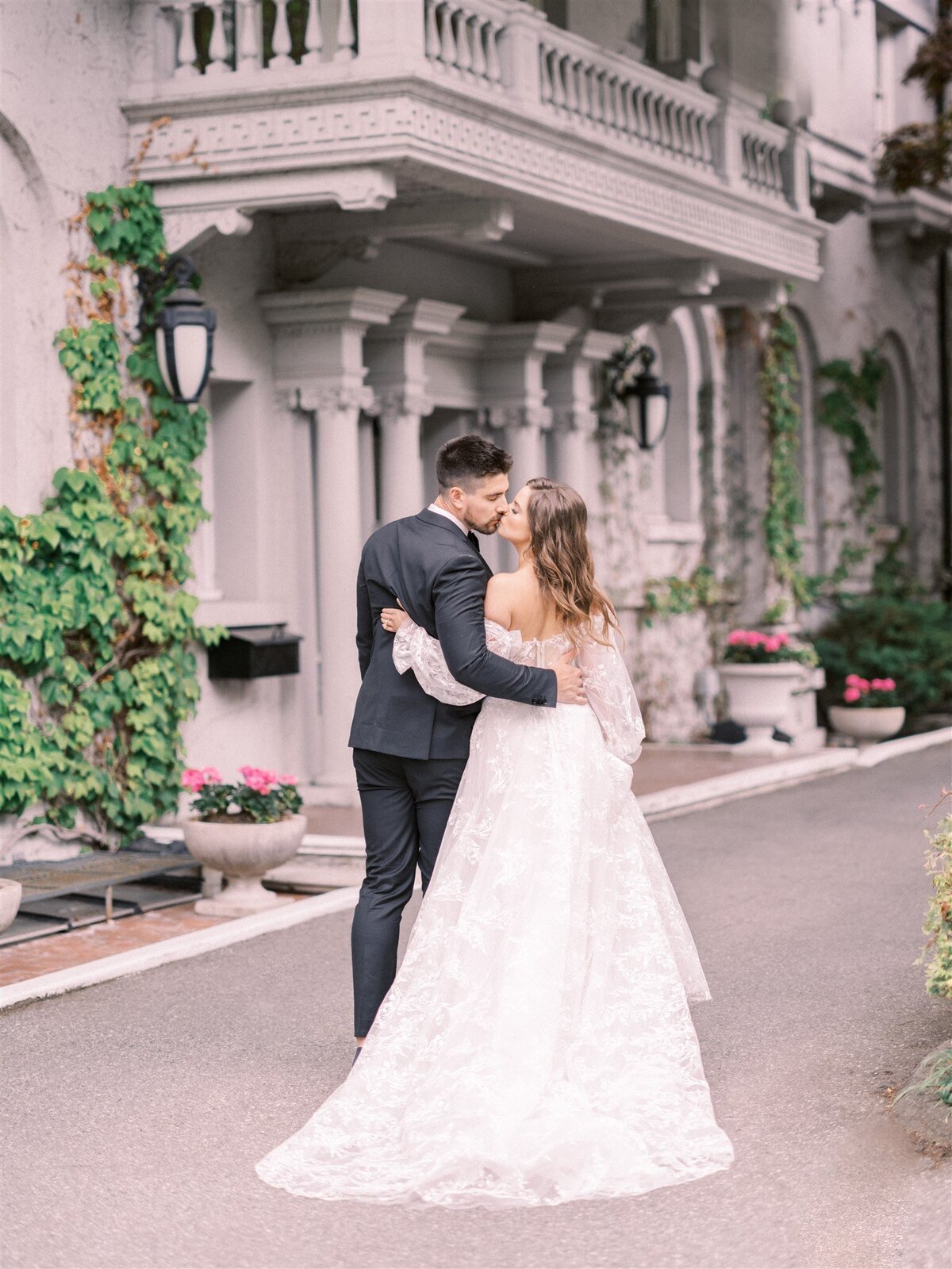 A couple in formal attire, with the woman in a white gown and the man in a dark suit, embrace on a driveway in front of a vine-covered building, showcasing their exquisite wedding design.