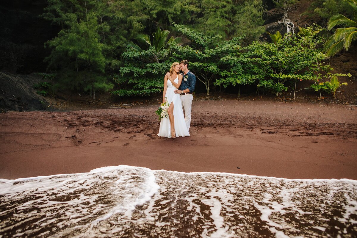 A bride and groom stand on the red sands beach during their adventure elopement in Hawaii
