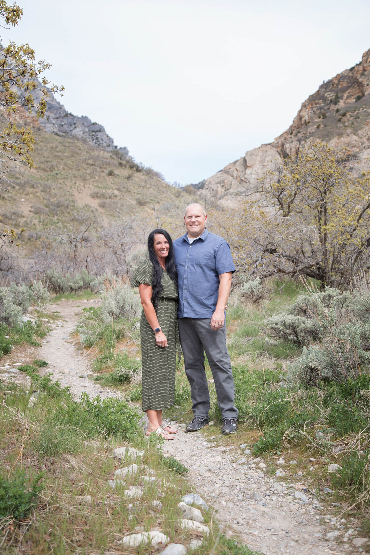 Woman with long black hair in green  romper standing by bald husband in blue shirt on mountain trail