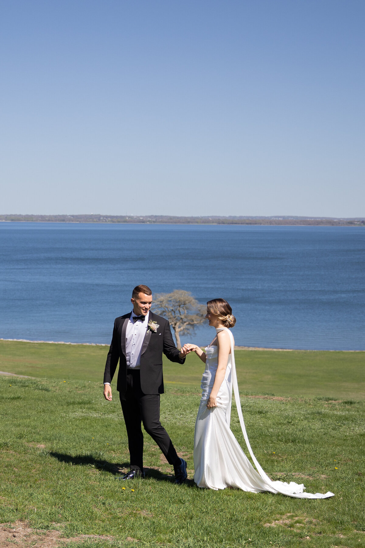 Aldrich Mansion Wedding Photos | Kelsey Sheehan Photography Timeless Rhode Island Weddings | A couple, dressed in wedding attire, joyfully walks hand in hand on a grassy hill. A vast, calm lake and clear blue sky form a serene backdrop.