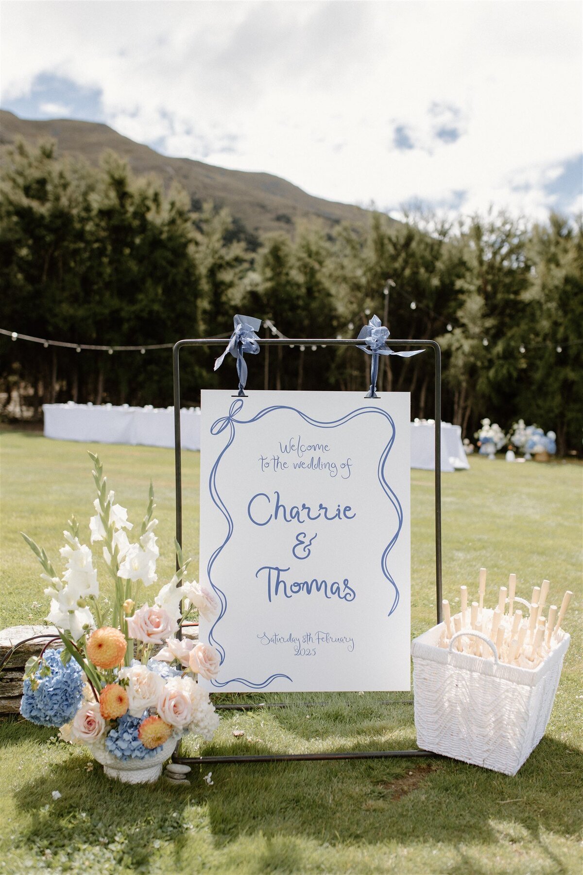 Outdoor wedding welcome sign for Charrie and Thomas, adorned with pastel flowers and parasols, set against lush greenery and hills.