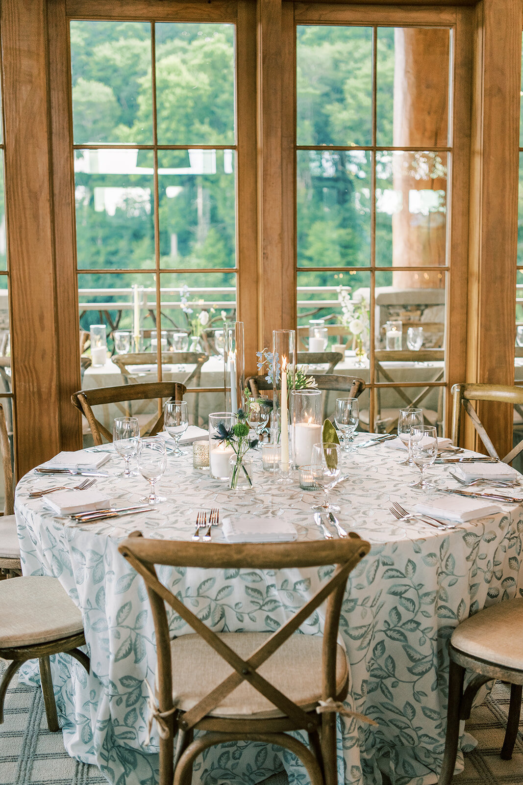 Round reception table with blue botanical linen, candles, and simple greenery centerpiece near large windows.