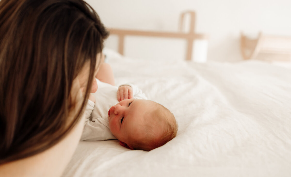 A mother lies on a bed in soft, natural light, dressed in cozy white clothes, as her newborn baby—snuggled in a white onesie—gazes up at her. This warm, intimate moment captures the quiet beauty of motherhood and the connection between mom and baby during an in-home newborn photography session in Maine.