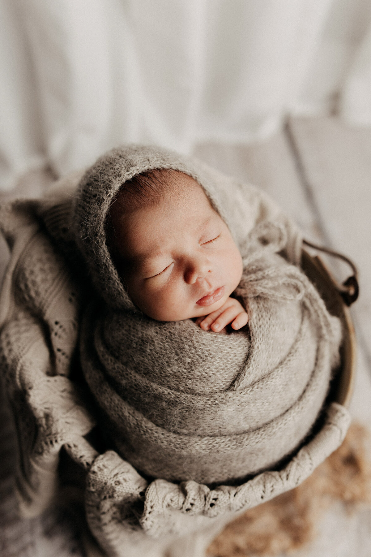 Newborn baby peacefully sleeping, swaddled in a soft taupe knit blanket with a matching bonnet, nestled in a cozy basket.