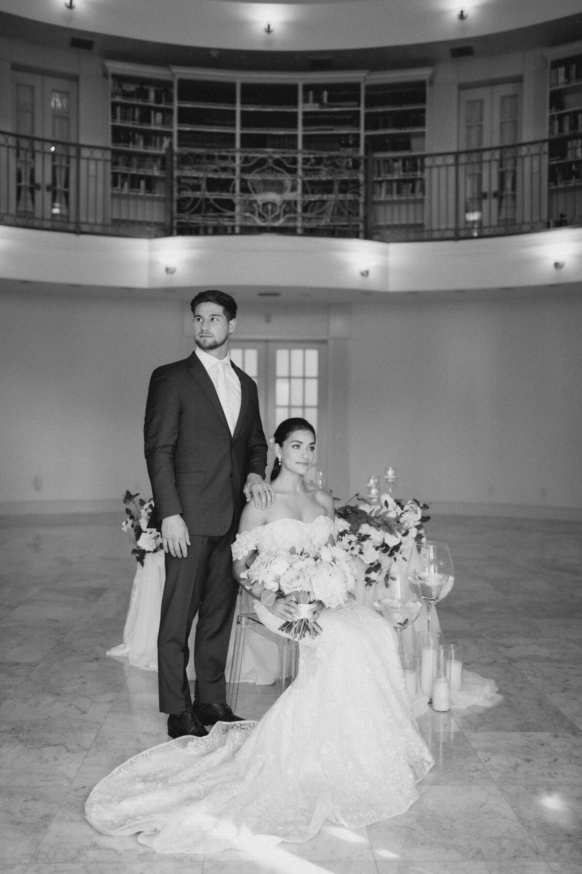 Black and white photo of bride and groom standing in front of an elegantly decorated sweetheart table, capturing a timeless and romantic wedding moment by In Full Bloom Event Planning & Design