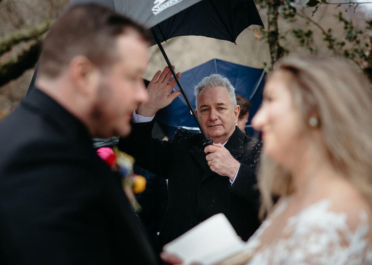 A groom smiling towards the bride with an officiant holding an umbrella in the background during a wedding ceremony