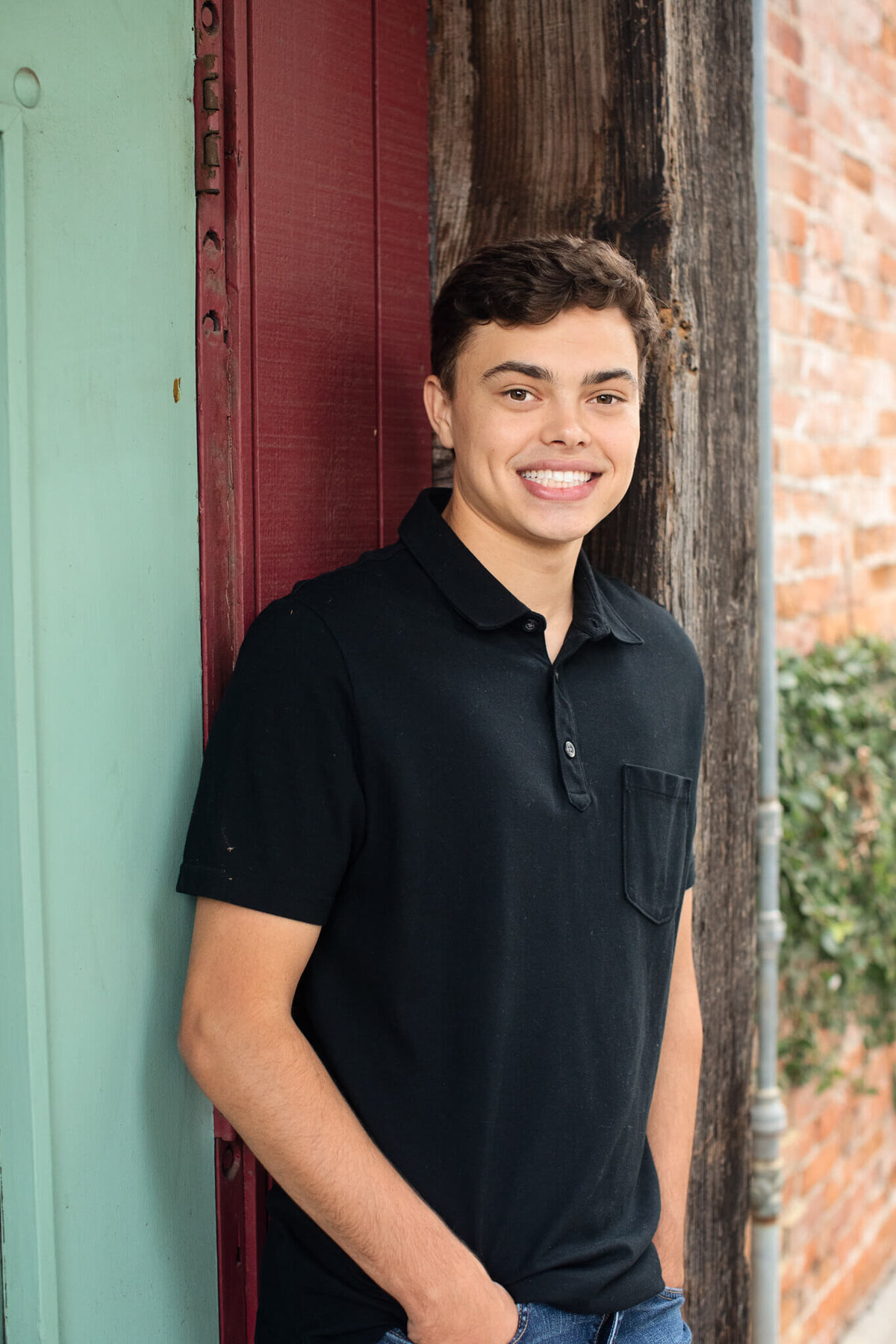 A young man in a black polo shirt stands beside a wooden and brick wall, smiling with hands in his jean pockets. The background includes a green door and an ivy-covered brick surface.