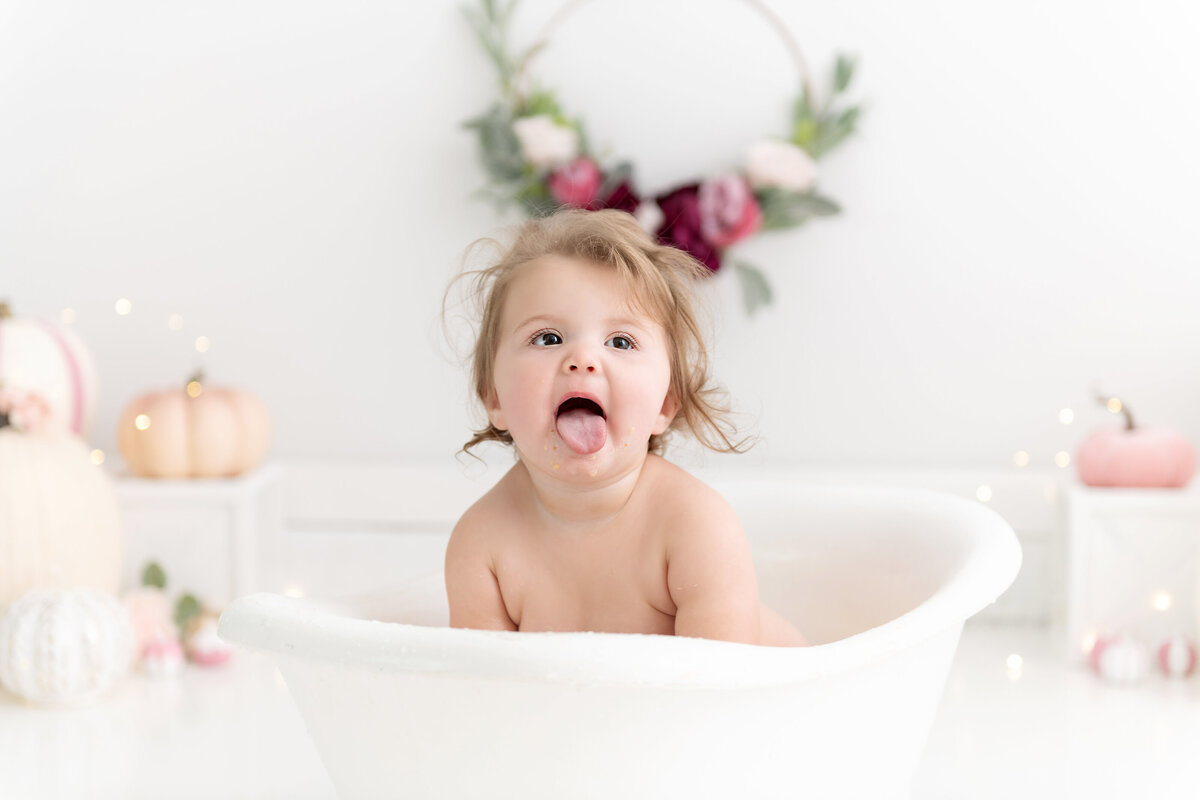 A toddler girl sits in a bathtub in a studio sticking out her tongue