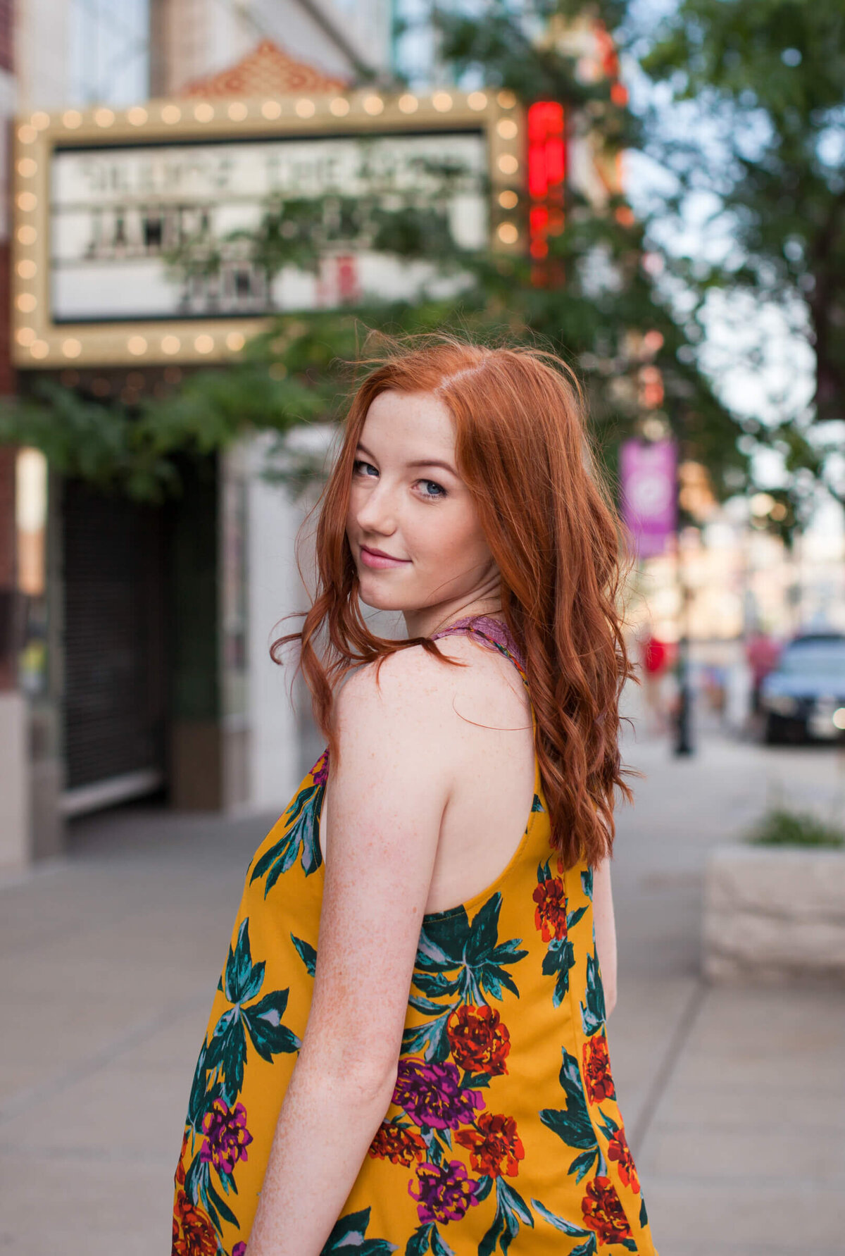 A lovely redhaired teen looks back over her shoulder with wind blown hair on a city street with a theater marquise in the background. Captured by Springfield, MO senior photographer Dynae Levingston.