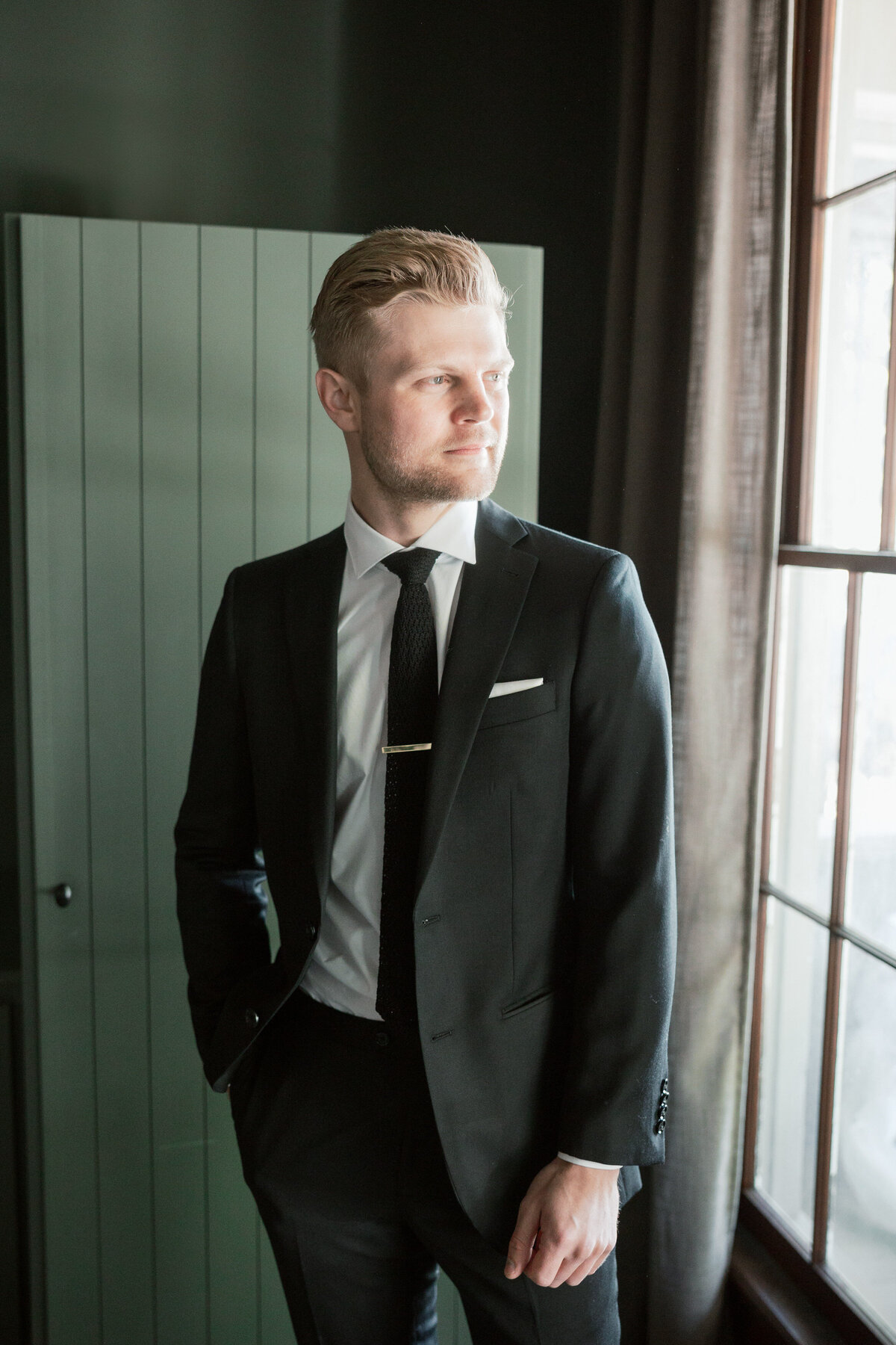 Groom in black suit stands by window with hands in pockets, thoughtful portrait by a Philadelphia wedding photographer in Dayton.