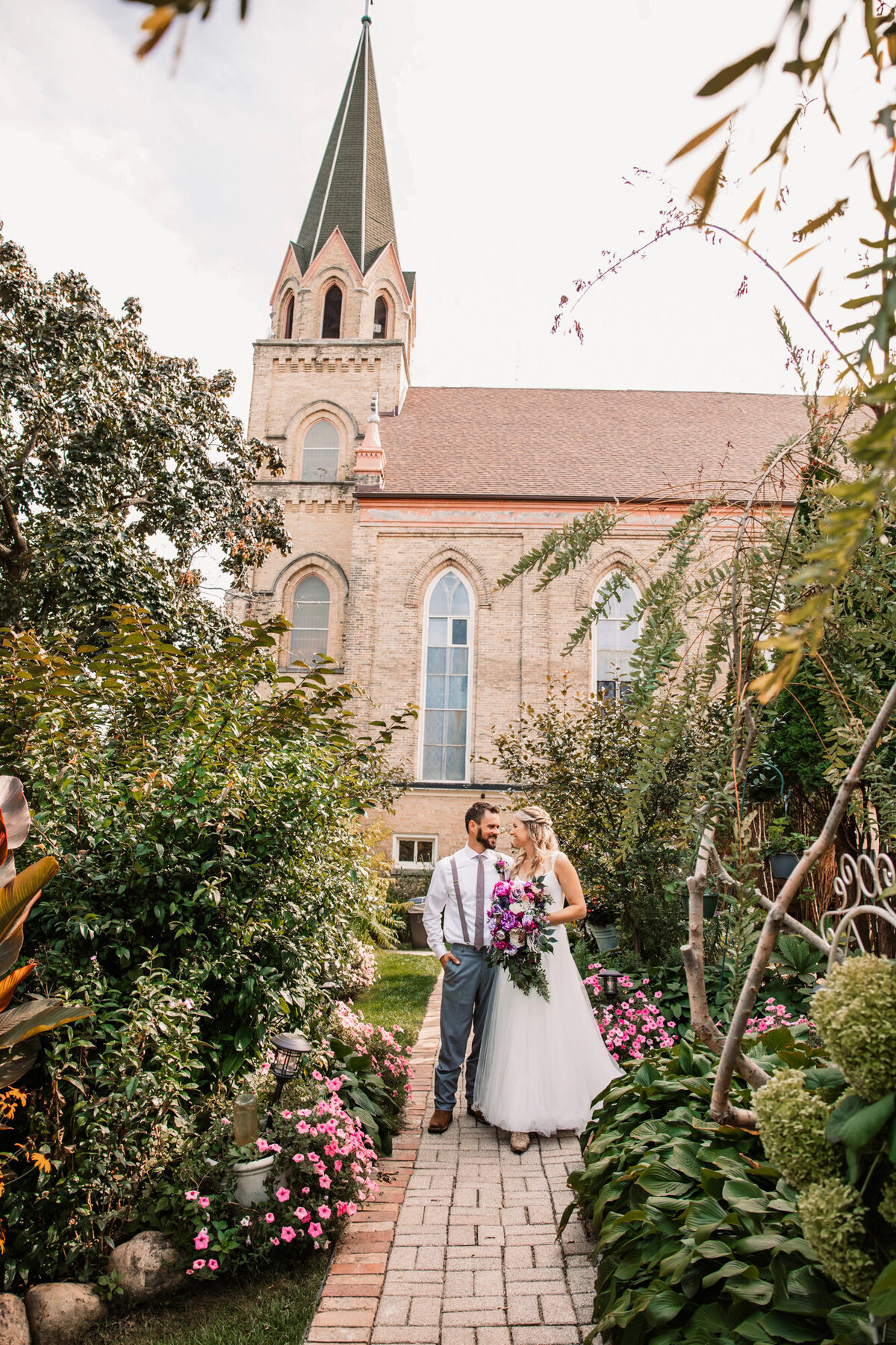 bride and groom stand on brick path in front of a church surrounded by lush gardens