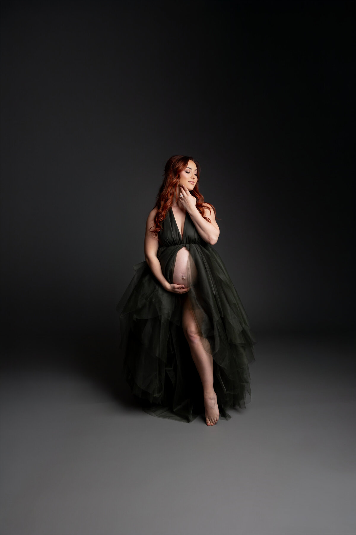 In a black dress, she tenderly holds her belly, her calm expression framed by a dark, dramatic backdrop. Captured by Inland Empire photographer Kaitlyn Dawn Photography.