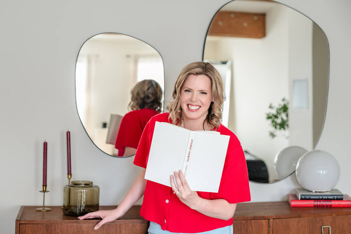 Brittany holding open book and smiling in red knit top.