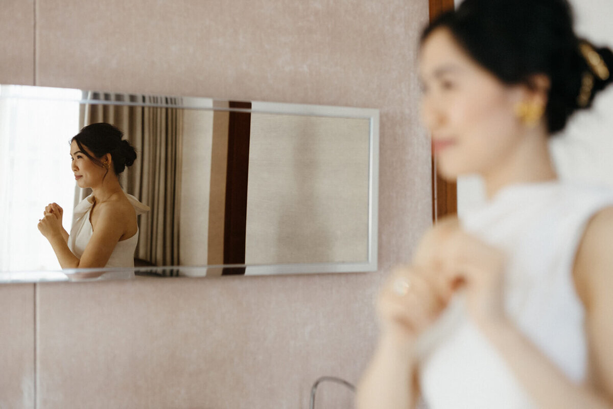 A bride's reflection in the mirror with her closed hands raised above her chest, and a blurred image of her facing the mirror