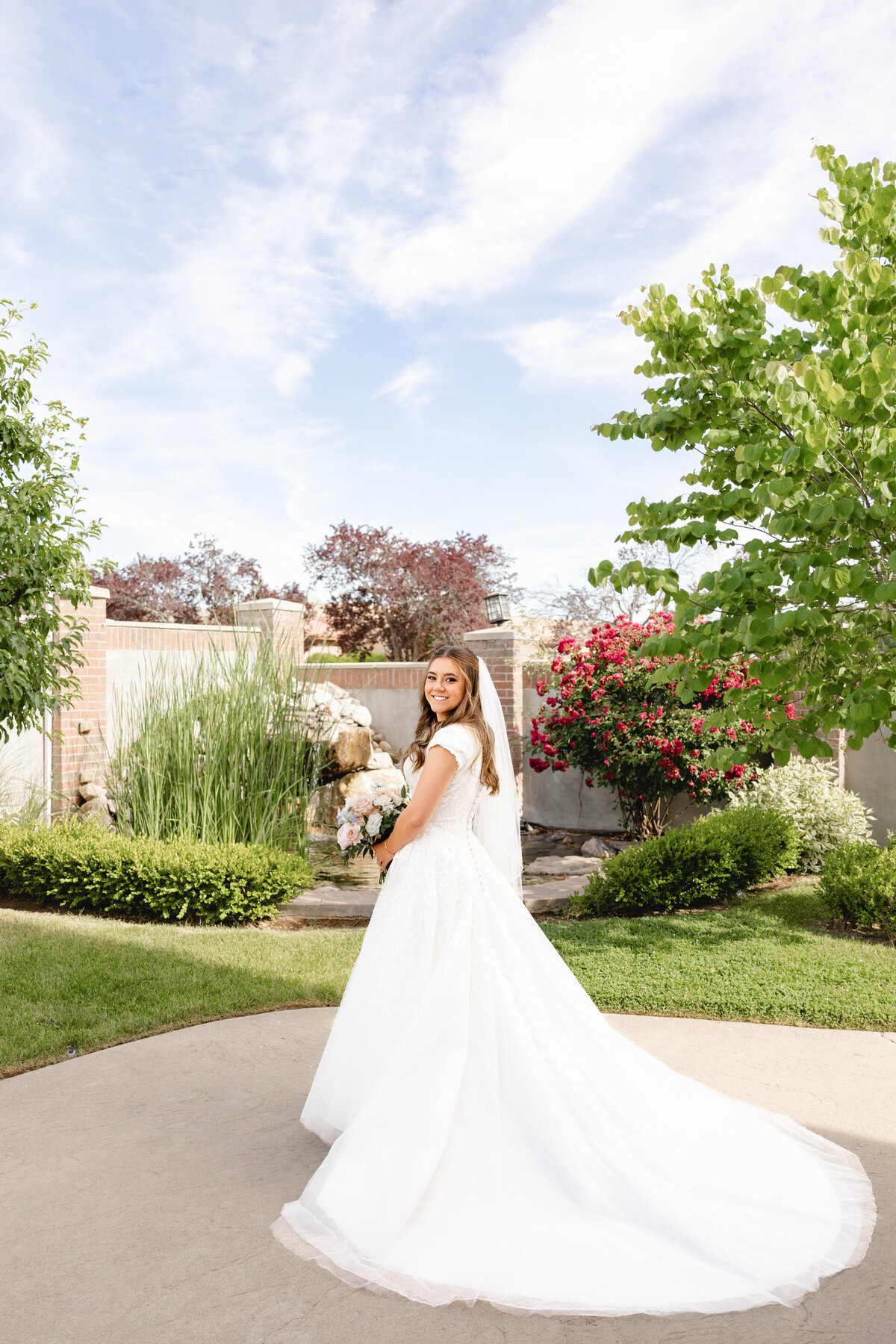 A radiant bride poses in a classic white wedding gown, standing in a beautifully landscaped garden with pink and green foliage.