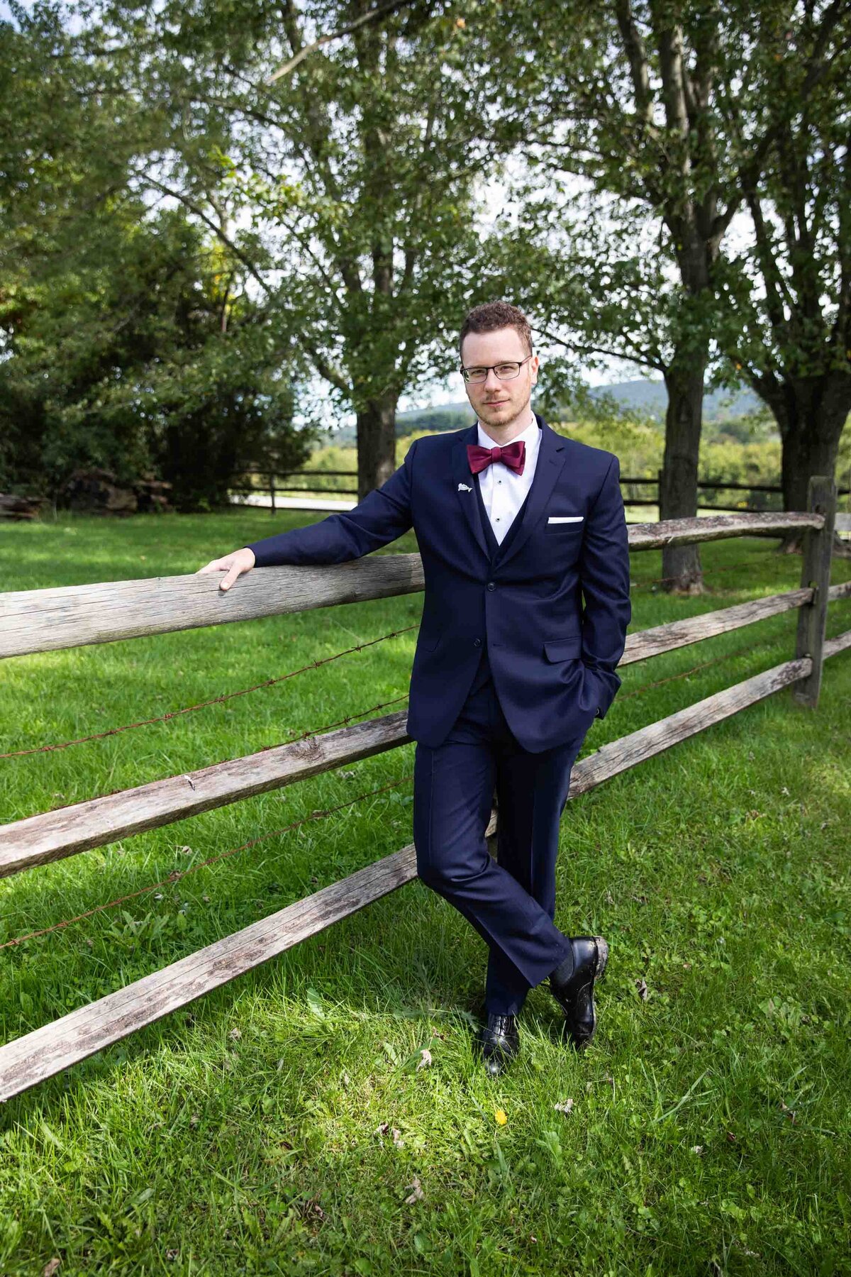 Portrait of a groom, standing in front of a wooden fence at Zigbone Farm Retreat, wearing a navy suite