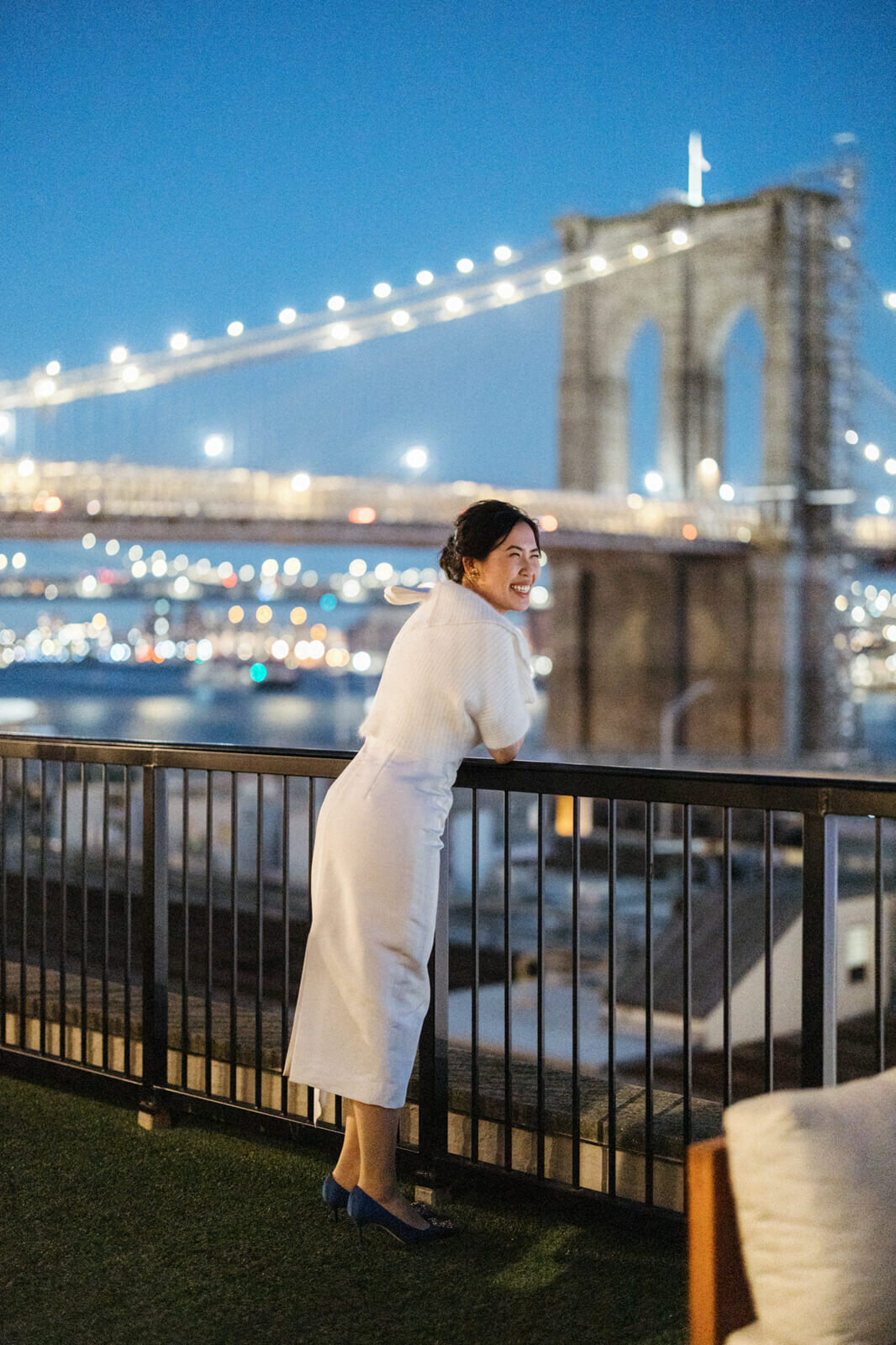 Side view of the smiling bride, leaning over a veranda railing, overlooking the Brooklyn Bridge in the night.
