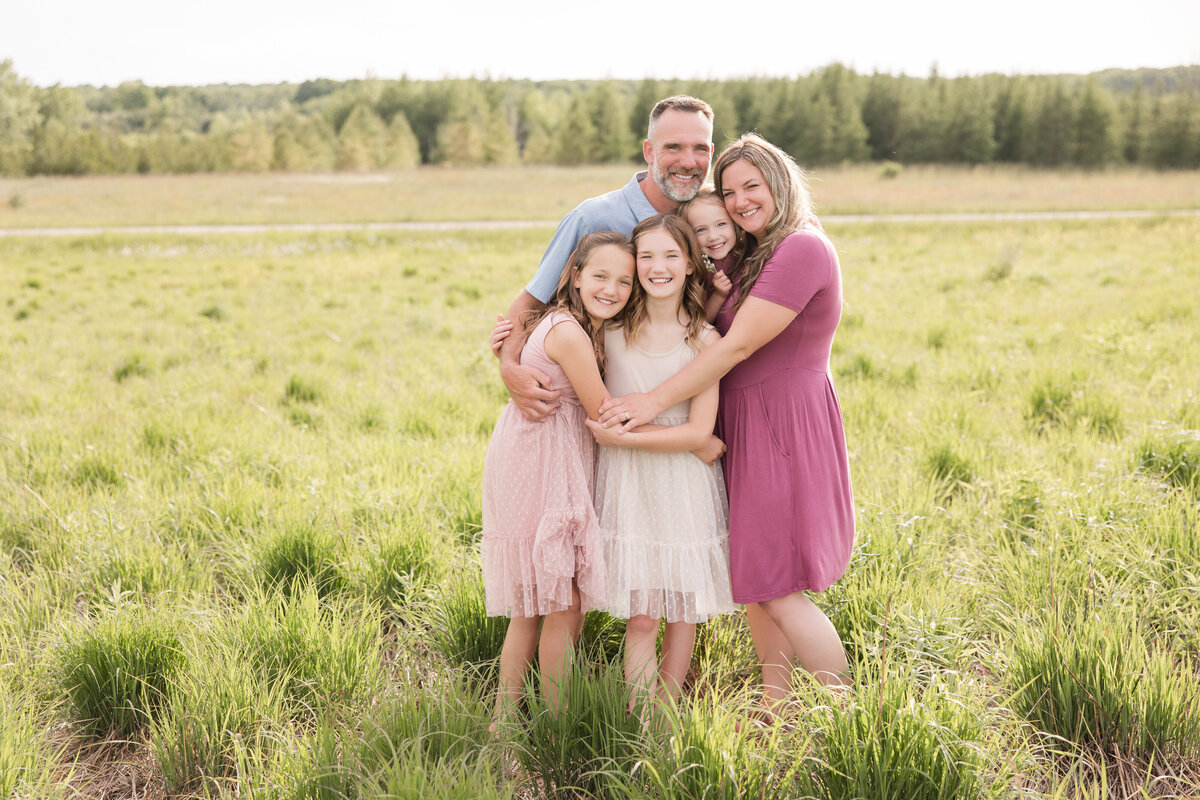 A family is embracing one another in a grassy field and smiling at the camera
