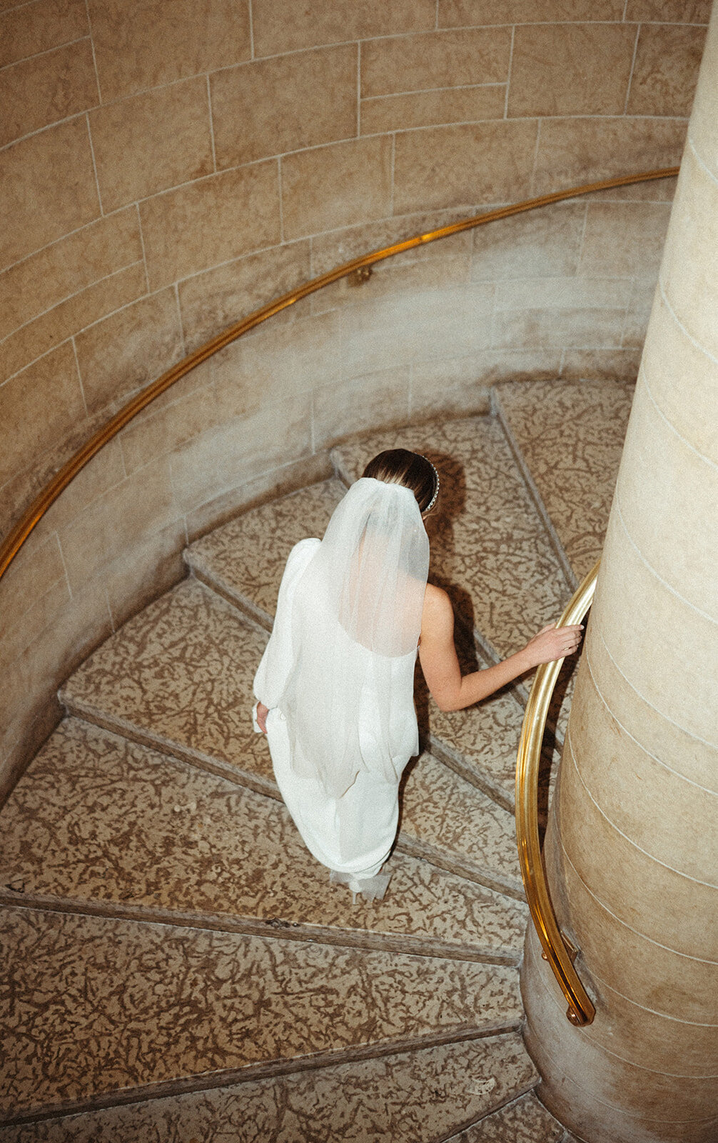 A bride in a white dress with a veil descends a curved staircase, holding onto a brass handrail. The staircase, designed by our full service wedding planner, features stone walls and ornate patterned steps.