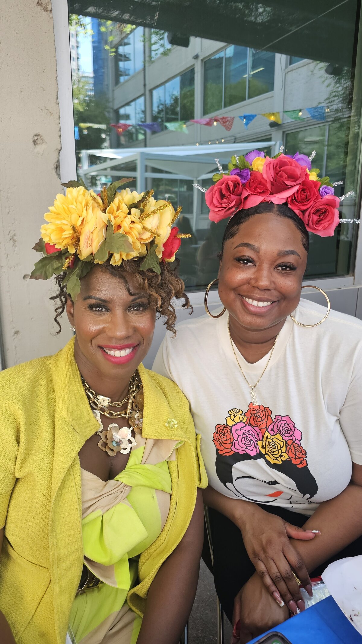 The beauty of floral elegance as two seated women showcase their artistic spirit adorned with handmade flower crowns in a vibrant colors. Dressed in stylish, artistically printed t-shirts and complemented with gold necklaces and earrings, these women embody the essence of creativity at Soleil Vida Studio