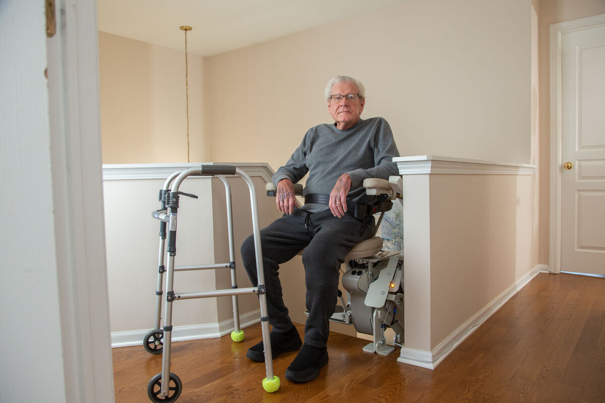 An elderly man sits in a chair with a walker next to him