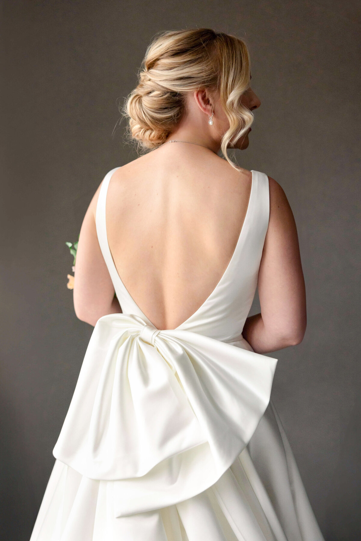 A woman in an elegant white wedding dress with a low back and large bow stands facing away. Her blonde hair is styled in an updo with loose curls. She holds a small bouquet in her left hand against a plain, dark background.