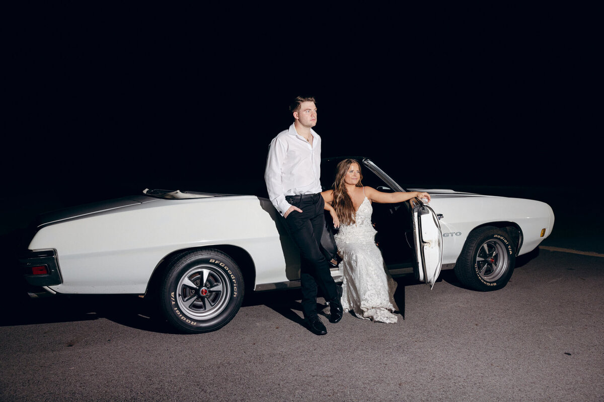 A wedding photo of the bride and groom posing with their getaway car at the end of the wedding reception