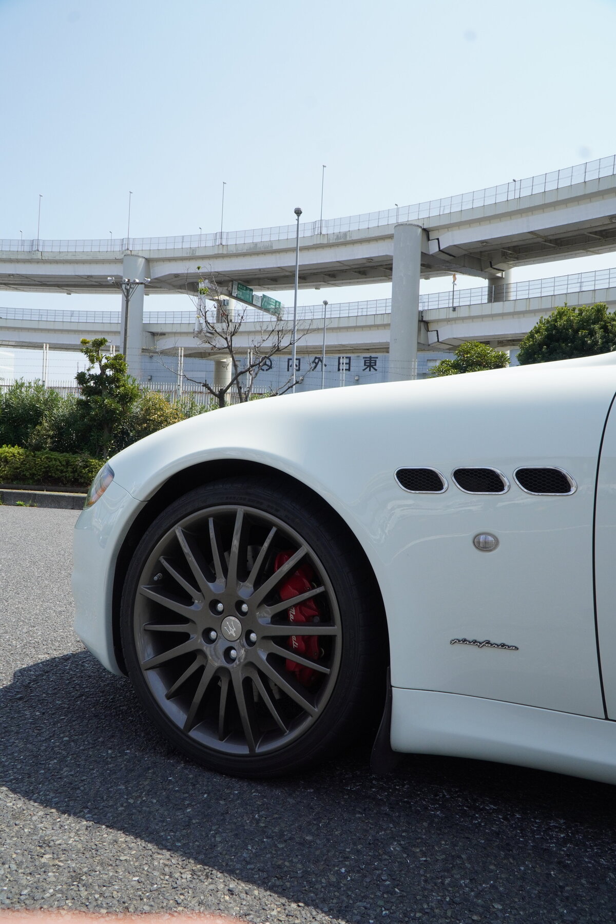 Close-up view of a white Maserati Quattroporte GTS showcasing its sporty front wheel, red brake calipers, and triple side vents with an elevated highway structure in the background.