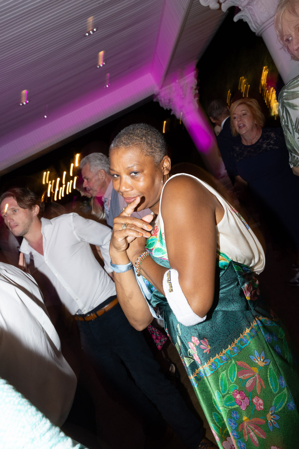 A woman posts for a photo at an indoor wedding reception at night. The people are her are slightly blurred.