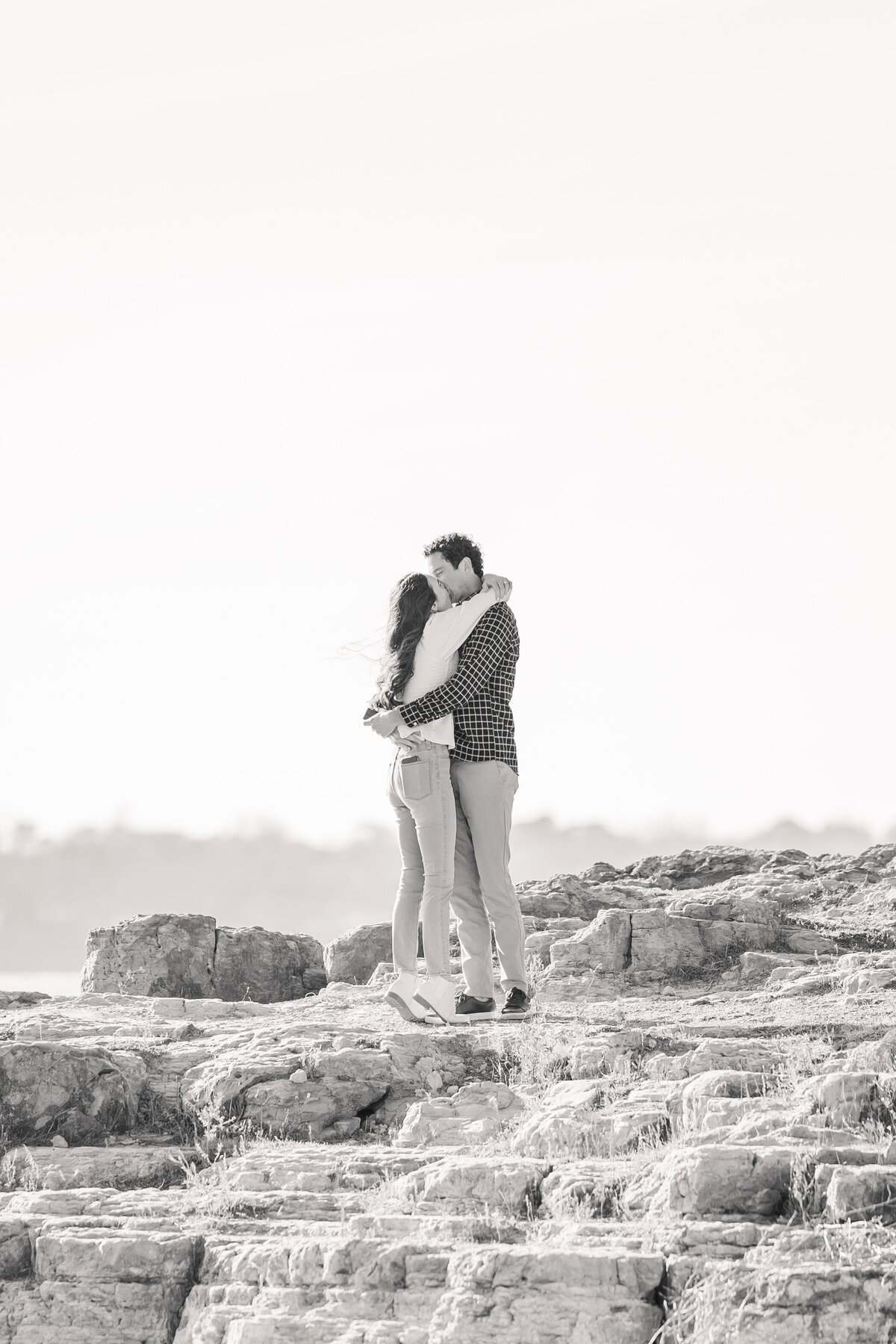 Timeless black and white photo of a romantic proposal at Lake Grapevine. A joyful couple shares an embrace on the rocks with the lake in the distance, capturing the emotion of the moment beautifully.