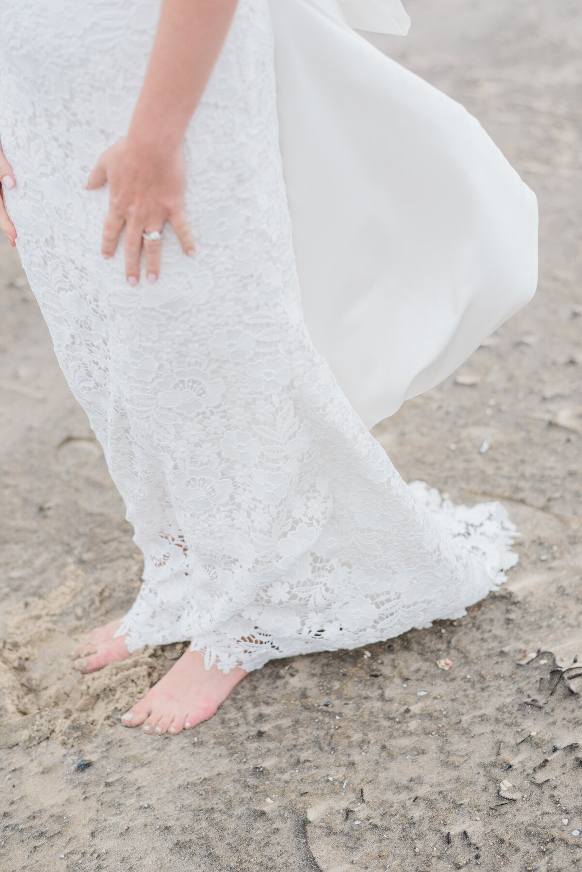 A close-up of the bride’s white lace wedding dress as she stands barefoot on the sandy beach.