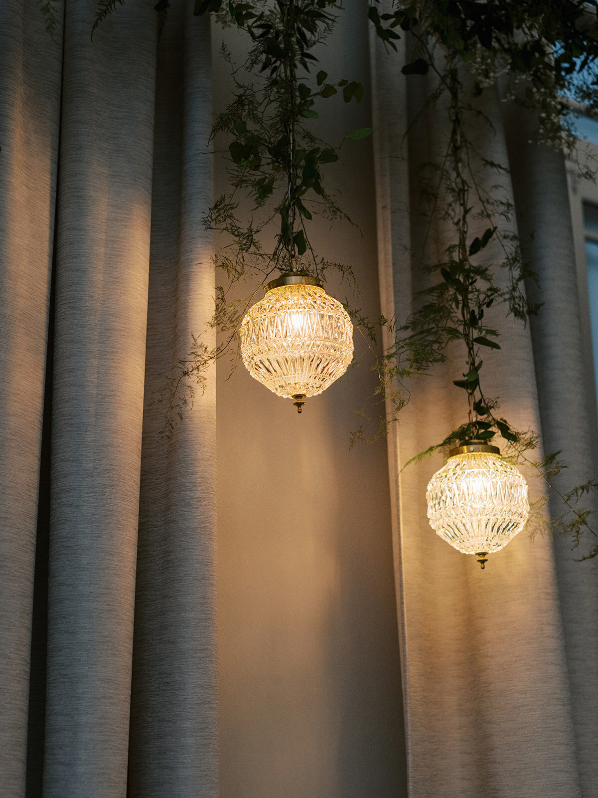 Two round crystal pendant lights are lit, hanging in front of light-colored curtains with some green foliage intertwined around them, creating an ambiance reminiscent of a classic Calgary wedding.