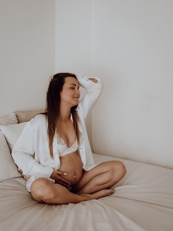 Een intiem portret van een zwangere vrouw die ontspannen glimlacht terwijl ze haar buik vasthoudt. Een moment van rust, vertrouwen en zelfliefde in een zachte setting.