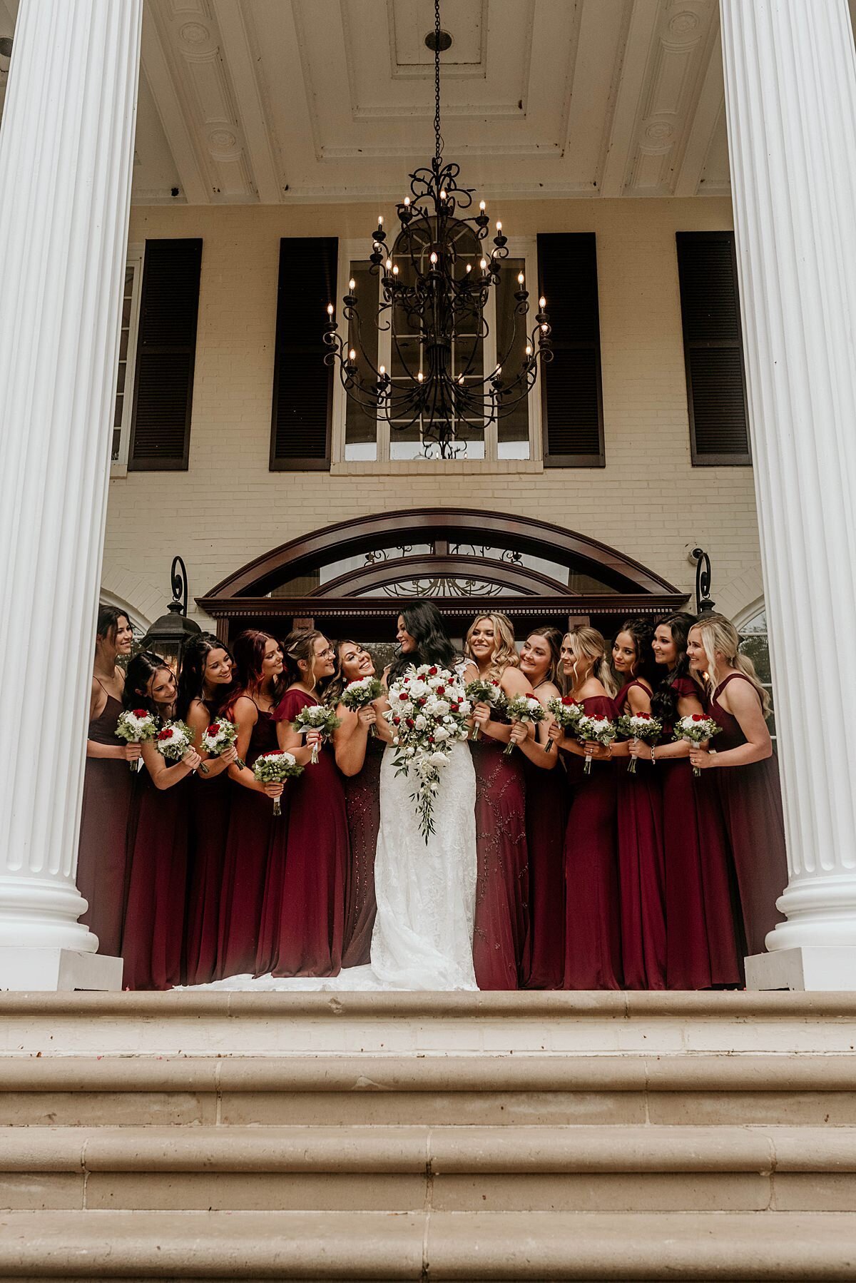 A bride, holding a large white cascade bouquet stands between her bridesmaids dressed in burgundy dresses as they stand on the steps of the mansion at The estate at Cherokee Dock.