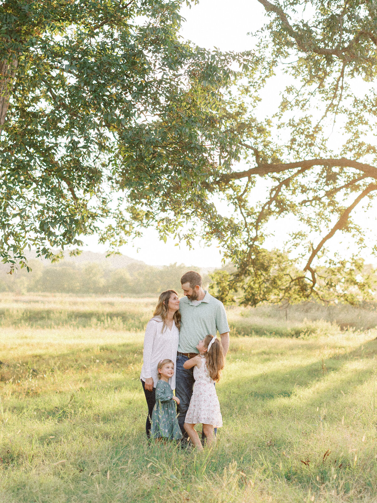 couple with two young girls stand close to one another underneath large tree