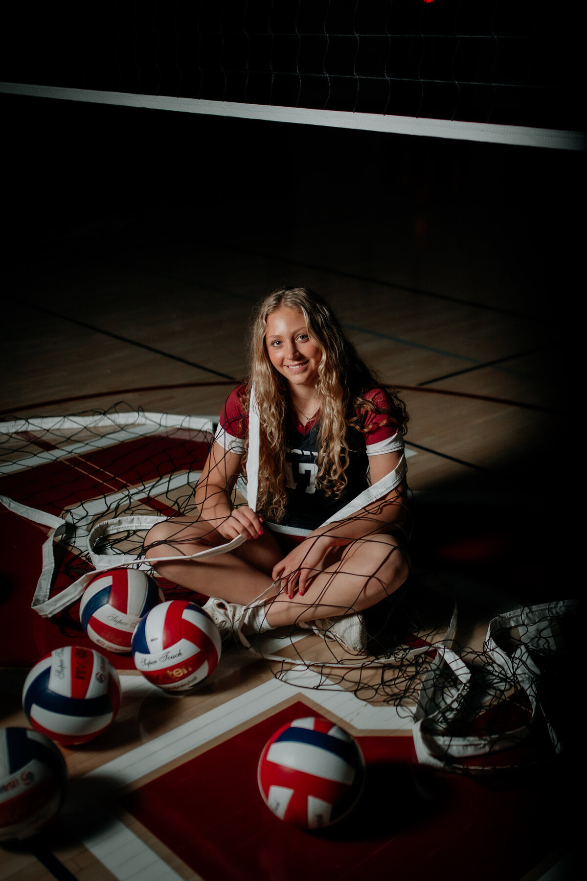 A senior volleyball player sits on the court, with a volleyball net draped around her during a sports themed senior session.