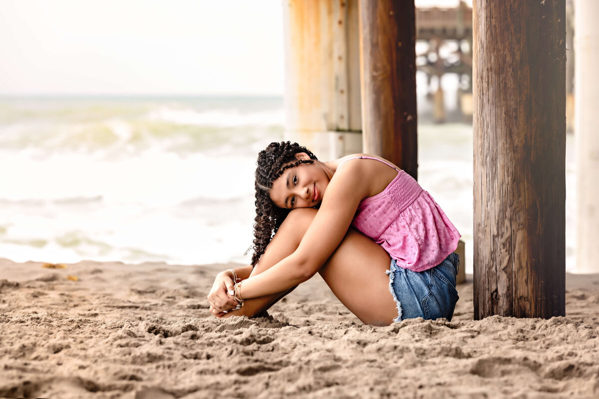 Cocoa Beach senior portraits under the pier with a side profile of a girl sitting in the sand, knees bent, serious expression and moody tones.
