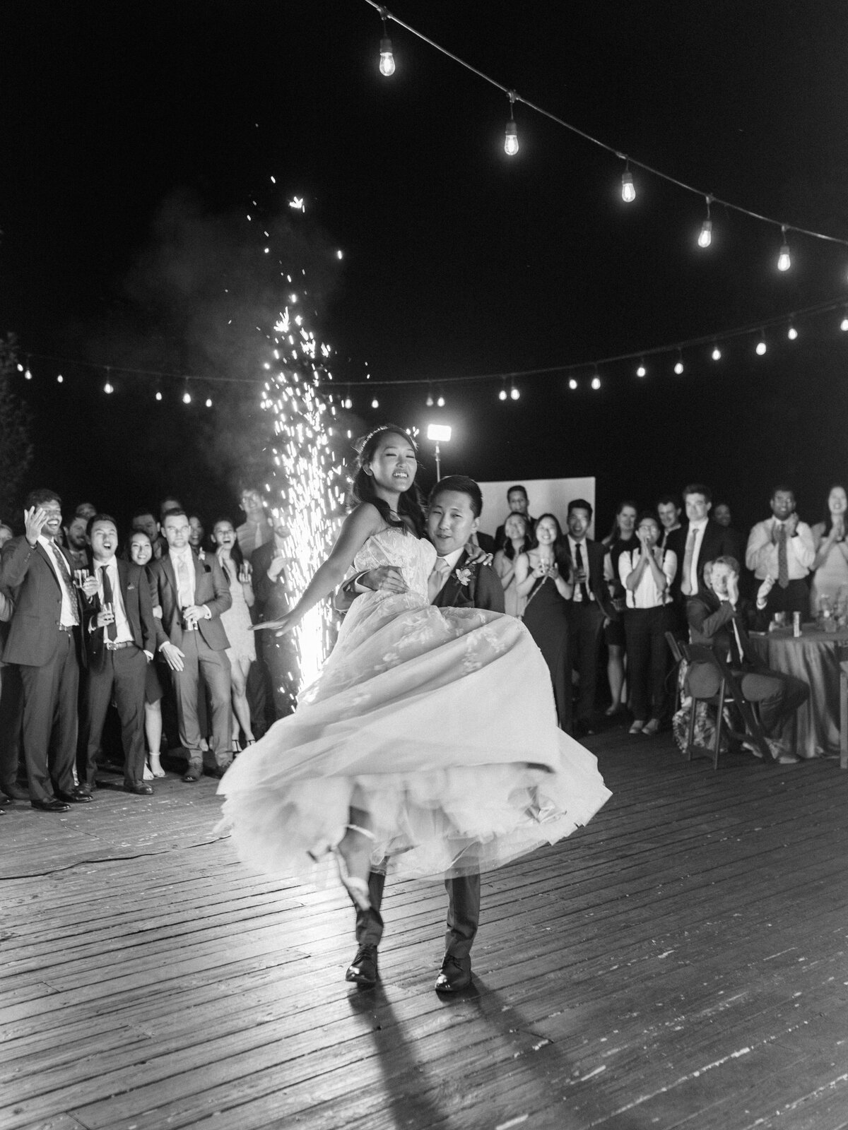 Black and white photo of bride twirling in her wedding gown during first dance as guests cheer in the background.